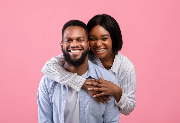 Портрет прекрасной черной женщины, обнимающей своего партнера сзади, улыбающейся и счастливой на розовом фоне студии - Фото, изображение