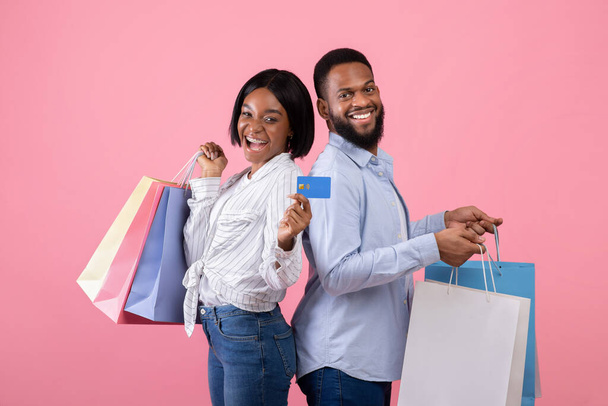 Portrait of black couple holding gift bags and credit card, shopping for Valentines gifts remotely on pink background - Photo, Image