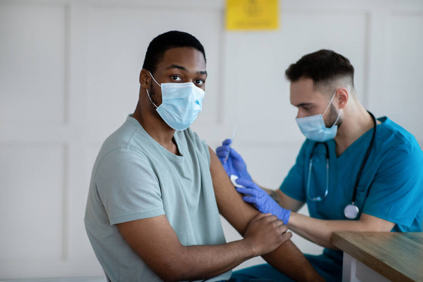 Trabajador de la salud en máscara quirúrgica que atiende a paciente varón negro, haciendo la inyección de la vacuna del coronavirus en la clínica - Foto, imagen