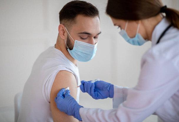 Vrouwelijke arts die een injectie met coronavirusvaccin toedient aan een jonge patiënt met een medisch masker in een gezondheidscentrum - Foto, afbeelding