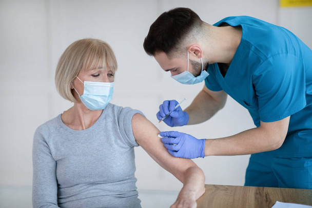 Maskeli kıdemli kadın Coronavirus 'a karşı aşı oluyor, tıbbi yardım alıyor, klinikte aşılanıyor. - Fotoğraf, Görsel