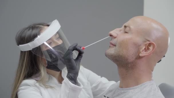 Test na stěr viru. Ženský doktor, který dělá nasofaryngeální stěr mužskému pacientovi. Lékař má na sobě lékařské ochranné prostředky: masku, ochranné brýle, rukavice a plášť. - Záběry, video
