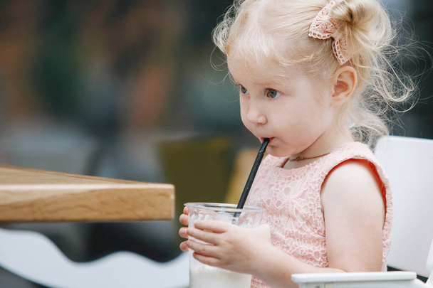 Blonde and a milkshake at a table on a city street. A three-year-old baby with ponytails in a pink dress poses emotionally for the camera. - Foto, imagen