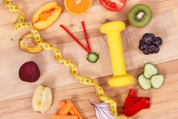 Fruits with vegetables in shape of clock, dumbbell for fitness and tape measure. Time to healthy lifestyle and nutrition concept - Foto, immagini