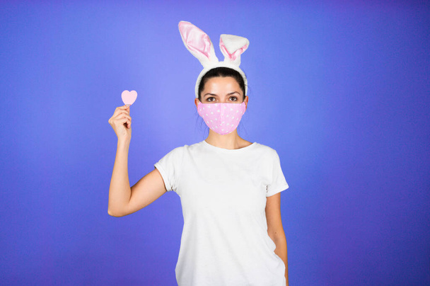 Jovem mulher vestindo impressão cardíaca máscara protetora e orelhas de coelho. Páscoas ou banner do dia de Velentine. Foto de alta qualidade - Foto, Imagem