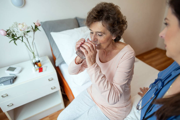 Senior Woman Patient Drinking Water. Elderly Female Patient Sitting on Hospital Bed with Nurse Watching Her. - Foto, imagen