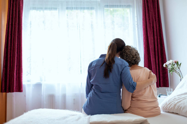 女性ホーム看護師ベッドの上で高齢女性の抱擁。高齢患者の肩の周りに彼女の腕を持つ女性看護師のバック妻. - 写真・画像
