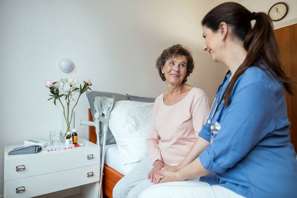 Mujer mayor y enfermera casera sentadas en la cama sonriéndose mutuamente. Paciente anciana alegre mirando al cuidador domiciliario en la habitación del hospital. - Foto, Imagen