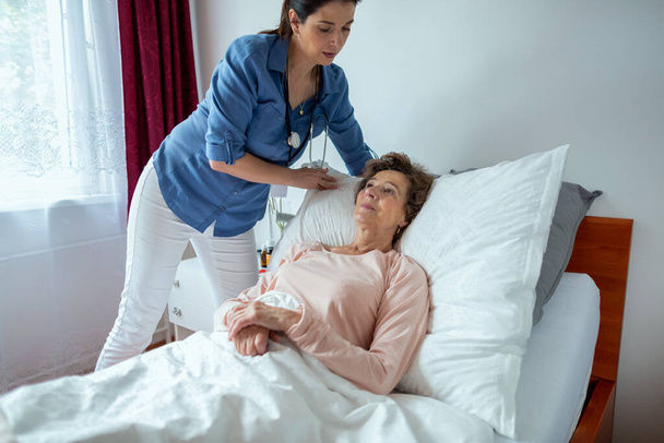 Home Nurse Making Elderly Patient Bed. Female Caregiver Adjusting Pillow For Senior Woman Lying in Hospital Bed. - Foto, afbeelding