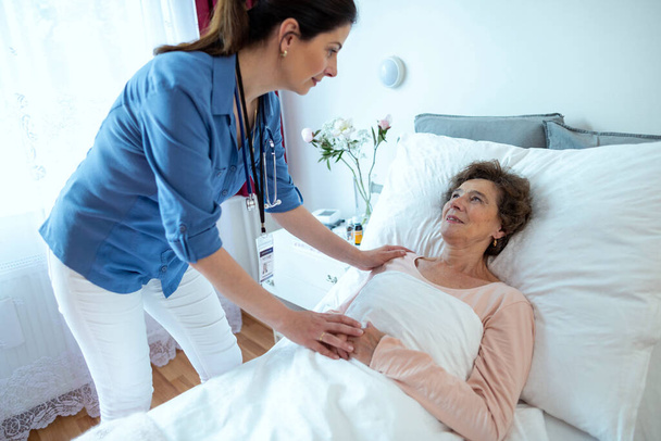 Infermiera appoggiata a una donna anziana sdraiata nel letto d'ospedale. Senior paziente femminile guardando a casa badante tenendo la mano. - Foto, immagini