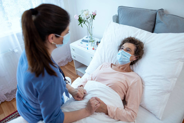 Пожилая женщина в защитной маске смотрит на медсестру, наклонившуюся. Top View of Home Caregiver with Face Mask Holding Hands of Female Senior Patient Lying in Bed at Nursing Home. - Фото, изображение