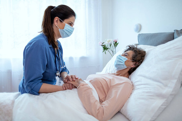 Утешитель дома Утешающий старшая пациентка лежит в постели. Вид сбоку на медсестру в маске хирургической защиты, разговаривающую с пожилой пациенткой в хосписе. - Фото, изображение