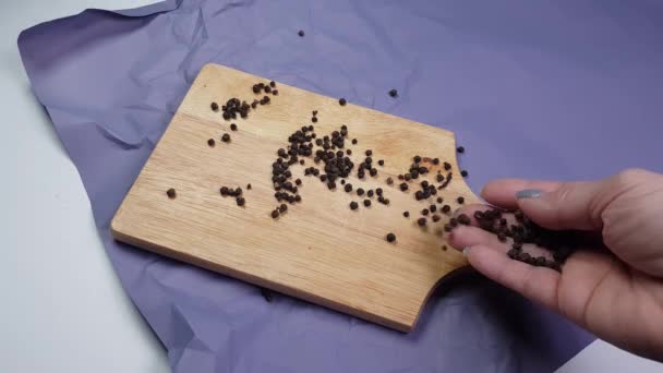 Piselli di pepe nero versati su un tagliere di legno, sdraiati su carta spiegazzata, una mano di donne versa spezie - Filmati, video