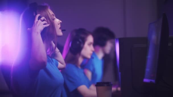 Grupa przyjaciół siedząca w klubie gier i mająca turniej gier online - kobieta zakładająca słuchawki i dołączająca do gry - Materiał filmowy, wideo