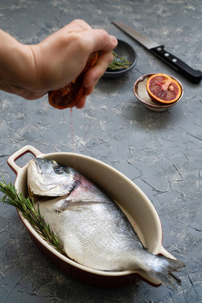 Μαγειρεύω θαλασσινά με εσπεριδοειδή και δενδρολίβανο. Η διαδικασία παρασκευής φρέσκων ψαριών για ψήσιμο ή τηγάνισμα με κόκκινα πορτοκάλια - Φωτογραφία, εικόνα