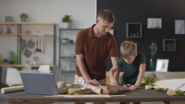 Medium slow-motion shot of young caucasian boy making flying kite cutting paper with construction knife and father helping him standing at table in living room - Footage, Video