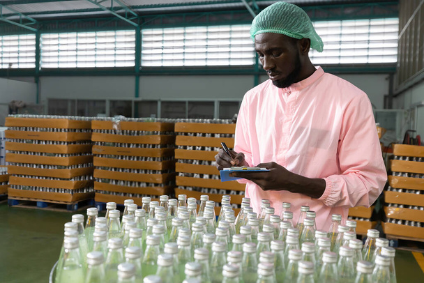 Trabajador africano de la fábrica que escribe en el portapapeles buscando y comprobando el inventario de productos en la fábrica de bebidas - Foto, Imagen
