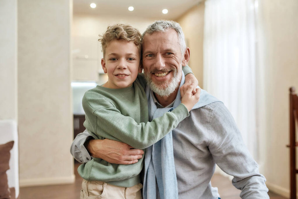 Happy grandfather embracing with his cute little preschool grandson, standing together at home and smiling at camera - Foto, imagen