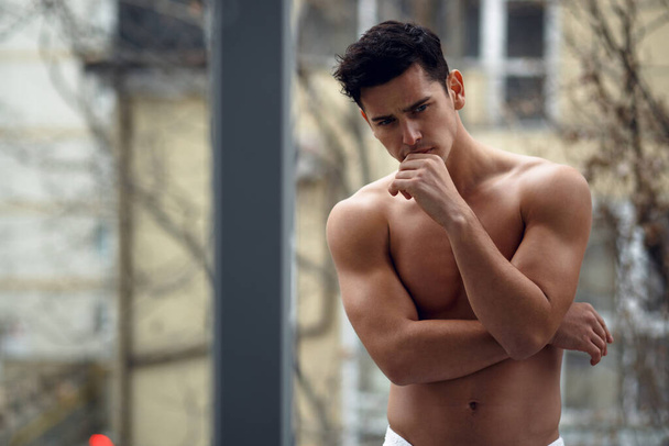 Hezký. bez košile svalnatý mladý muž v koupelně ručník, stojící na balkóně, izolované na ulici pozadí. - Fotografie, Obrázek
