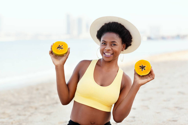 A young African girl in a summer straw hat is sitting on the beach, holding a papaya and smiling - Photo, image