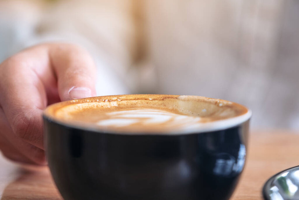 Closeup image of a hand holding a cup of hot coffee with latte art on wooden table - Valokuva, kuva