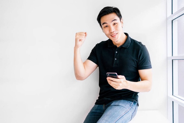 Good looking Asian male wearing a casual look at the camera while using a mobile phone feels victory and success, sitting near the window with natural light. Modern technology supports the business. - Photo, Image