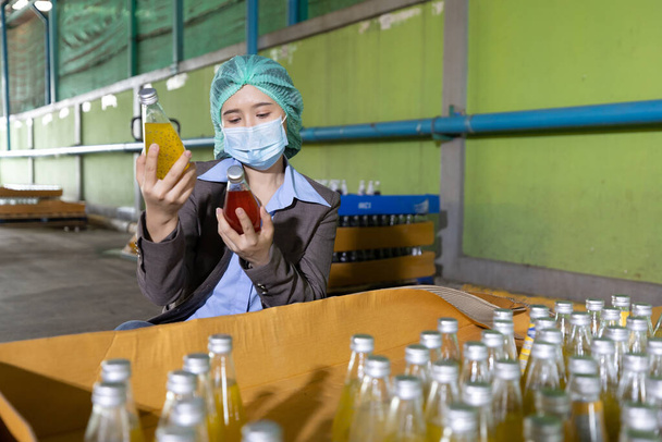 young woman manager wearing safety hat and medical mask looking basil seed drink for stocktaking of products in beverage factory - Photo, Image