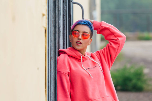 Young girl with short colorful hair and nose piercing dressed in pink hoodie and pink sunglasses satanding near the wall with wooden door. Copy space. - Φωτογραφία, εικόνα