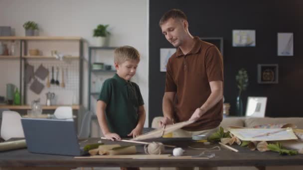 Medium schot van vader en zoon samen tijd doorbrengen crafting helder vliegend papier vlieger staan aan tafel in de woonkamer - Video
