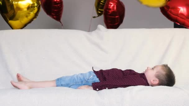 Sevgililer Günü için video görüntüleri. Eğlenceli bir parti. Beyaz bir çocuk, bir çocuk, hafif bir kanepede uzanıyor ve kalp şeklinde bir balonla oynuyor. Yavaş çekim - Video, Çekim
