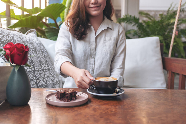 Wizerunek z bliska pięknej kobiety trzymającej i pijącej gorącą kawę latte z ciastem brownie na stole w kawiarni - Zdjęcie, obraz