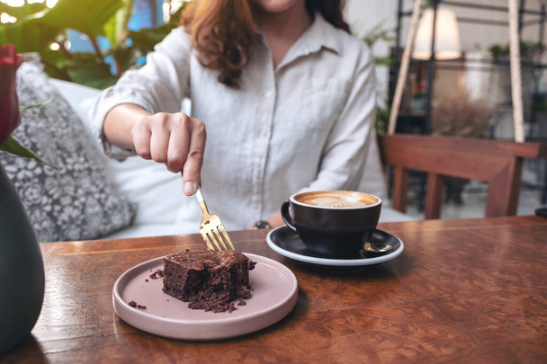 女性は、カフェで木製のテーブルの上にコーヒーカップとフォークでブラウニーケーキを切断 - 写真・画像