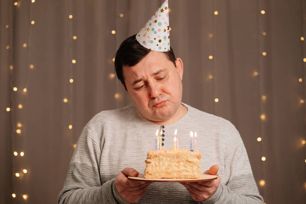 sad man in festive cap with birthday cake blows out candles.sadness due to aging - 写真・画像