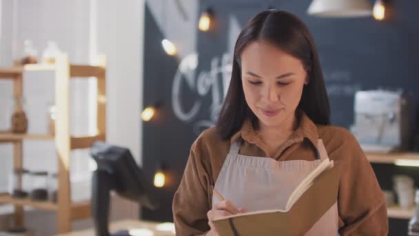 Mittlere Nahaufnahme Porträt einer jungen lächelnden asiatischen Frau, die ein kleines Café besitzt, Notizen in ihrem Notizbuch macht und dann lächelnd in die Kamera schaut - Filmmaterial, Video