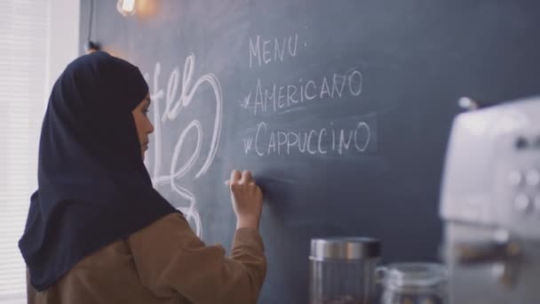 Μεσαία πλαϊνή άποψη γκρο πλαν του νεαρού μουσουλμάνου εργαζόμενου στο καφέ hijab γράφοντας ποτά καφέ στον μαυροπίνακα για τους πελάτες - Πλάνα, βίντεο