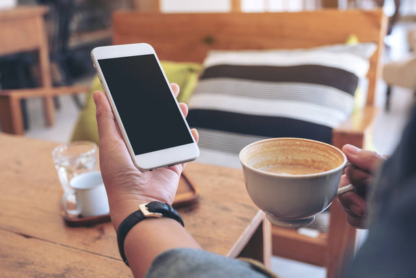 Imagen burlona de manos sosteniendo teléfono móvil blanco con pantalla en blanco mientras bebe café en la cafetería - Foto, Imagen