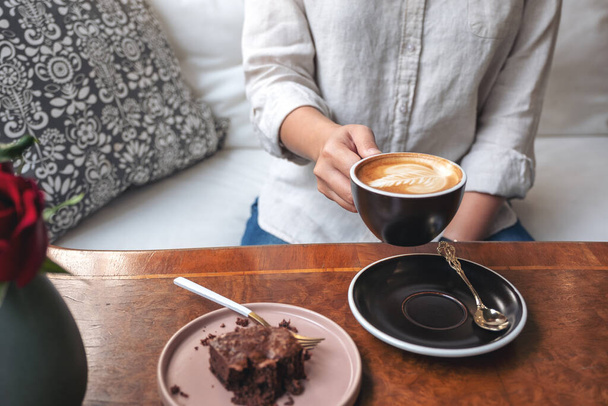 Closeup image of a woman holding and drinking hot latte coffee with brownie cake on the table - Zdjęcie, obraz