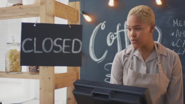 Запись юного афроамериканского продавца кофе в фартуке, стоящего у кассы и открывающего вывеску - Кадры, видео