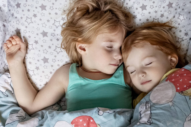 Δύο αδερφούλες αδελφές κοιμούνται σε μια αγκαλιά στο κρεβάτι κάτω από μια κουβέρτα. - Φωτογραφία, εικόνα