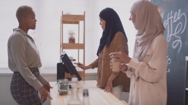 Imágenes medianas de una joven y alegre mujer afroamericana comprando café para llevar en una pequeña cafetería cubierta con dos trabajadoras musulmanas sonrientes en el mostrador - Metraje, vídeo