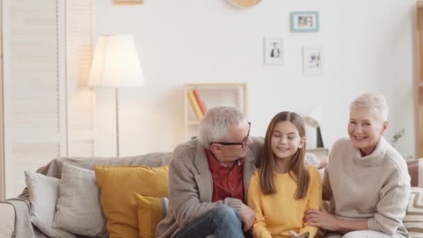 Aufgeweckte Aufnahme einer jungen kaukasischen Enkelin, die zu Hause mit Großmutter und Großvater auf dem Sofa sitzt, redet, sich dann alle umdreht, in die Kamera schaut und lächelt - Filmmaterial, Video