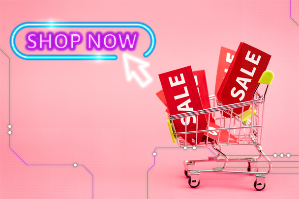 red labels with sale in small shopping trolley near shop now lettering on pink - Photo, Image
