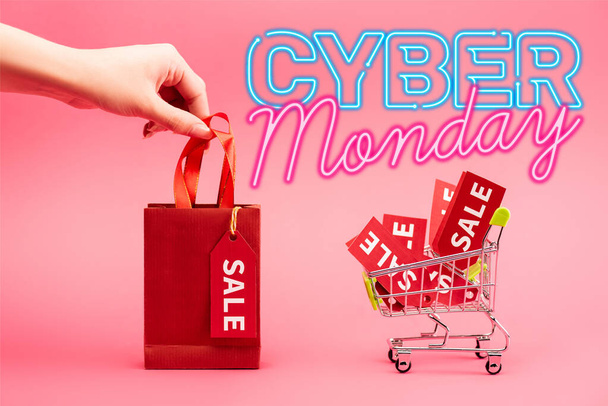 cropped view of woman holding small shopping bag near cart with sale tags near cyber monday lettering on pink - Zdjęcie, obraz