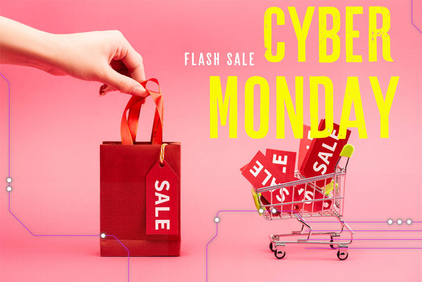 vista recortada de la mujer sosteniendo una pequeña bolsa de compras cerca del carrito con etiquetas de venta cerca de letras de lunes cibernético en rosa - Foto, imagen