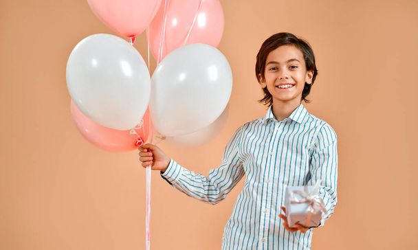 Cute caucasian smiling boy in a striped shirt, holding balloons on a background of powdery peach and a gift box. Valentine's Day. - Foto, Bild