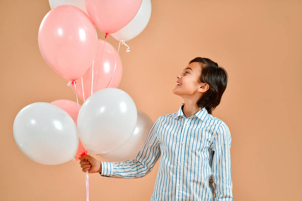 Miły biały uśmiechnięty chłopiec w pasiastej koszuli trzymający balony z helem odizolowanym na pudrowym brzoskwiniowym tle.. - Zdjęcie, obraz