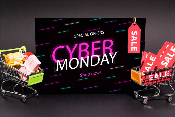 small presents in toy shopping carts near placard with special offers, cyber monday, shop now lettering on dark background - Photo, Image