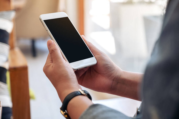 Imagen burlona de una mano sosteniendo teléfono móvil blanco con pantalla en blanco con taza de café en la mesa de madera en la cafetería - Foto, Imagen