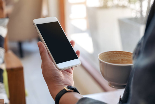 Imagen burlona de manos sosteniendo teléfono móvil blanco con pantalla en blanco mientras bebe café  - Foto, imagen