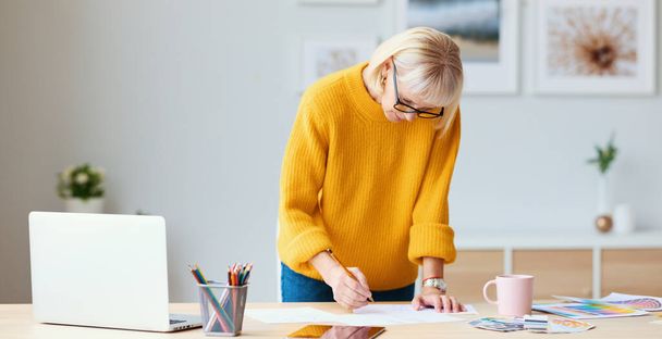 Seniorin in Pullover und Brille lehnt am Tisch und skizziert auf Papierbogen, während sie an einem Designprojekt im kreativen Arbeitsraum arbeitet - Foto, Bild